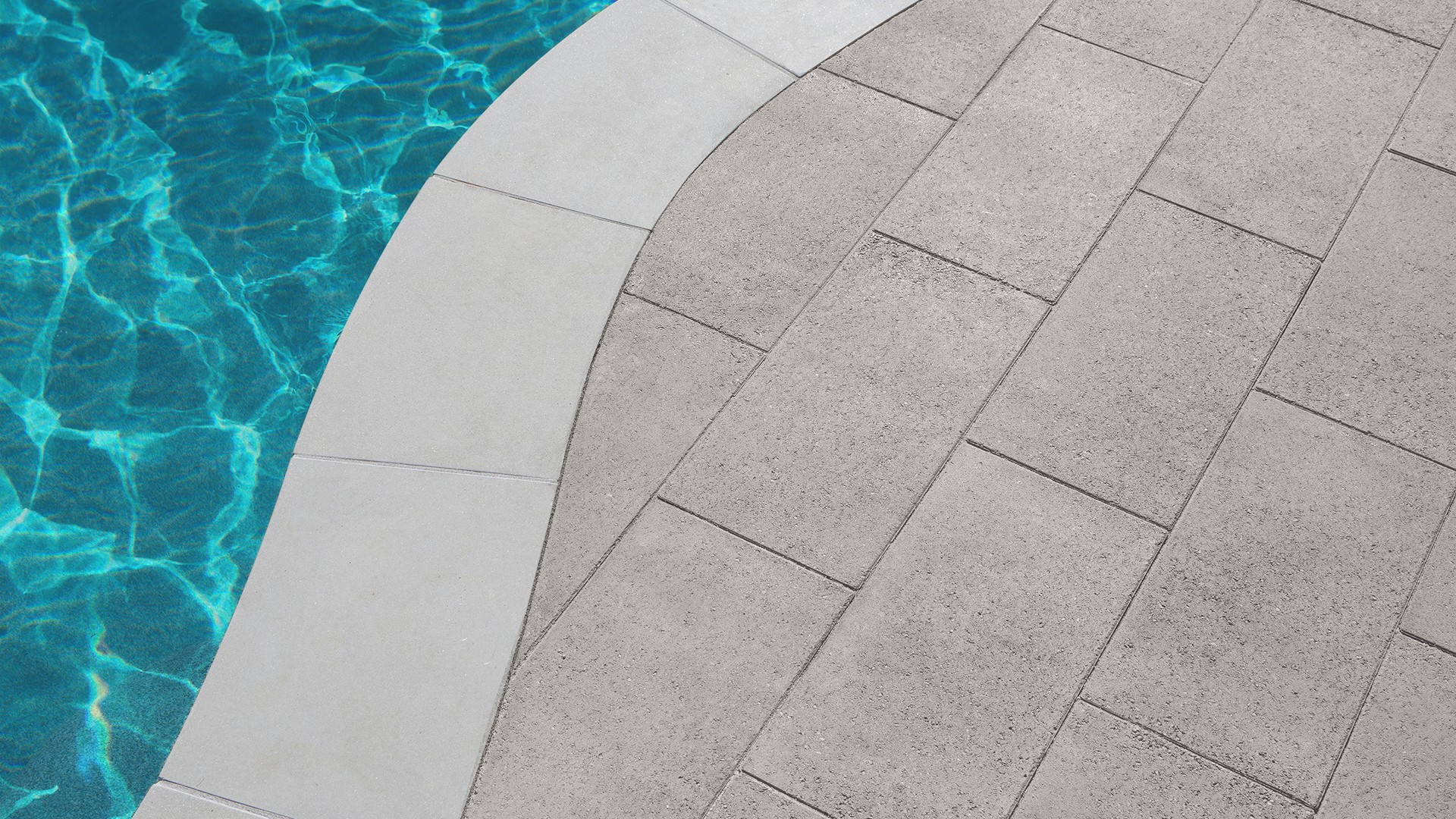 Close-up view of a poolside patio featuring 60mm 12x24 pavers in Solid Gray, installed in a running bond pattern. The curved pool coping aligns seamlessly with the edge of the water, creating a smooth transition between the textured pavers and the glistening blue pool surface. The overall design showcases clean lines and modern simplicity.