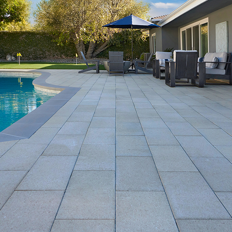 Modern backyard patio with 60mm 12x24 pavers in Solid Gray installed in a running bond pattern. The clean-lined pavers surround a curved swimming pool, which features darker gray coping. The poolside area is furnished with cushioned outdoor lounge chairs and a table under a navy umbrella. A neatly maintained lawn and mature trees complete the tranquil residential setting.