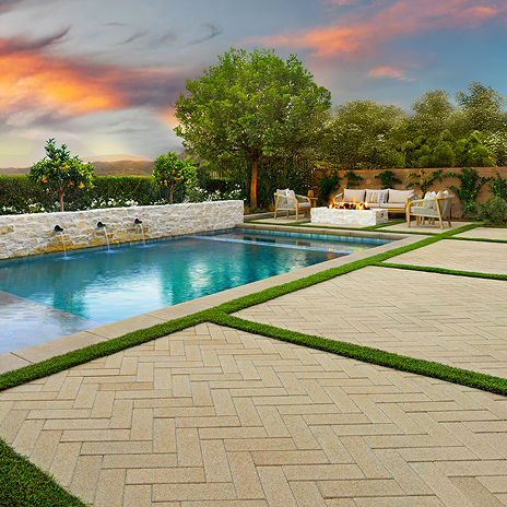 Elegant backyard pool area featuring 80mm 4x12 pavers in Light Sand, installed in a herringbone pattern. The pavers are arranged in clean geometric sections divided by green grass strips. The design surrounds a rectangular pool with water features flowing from a white stone wall, and a cozy fire pit seating area with cushioned chairs under a large tree. Lush landscaping and a vibrant sunset sky complete the tranquil outdoor setting.