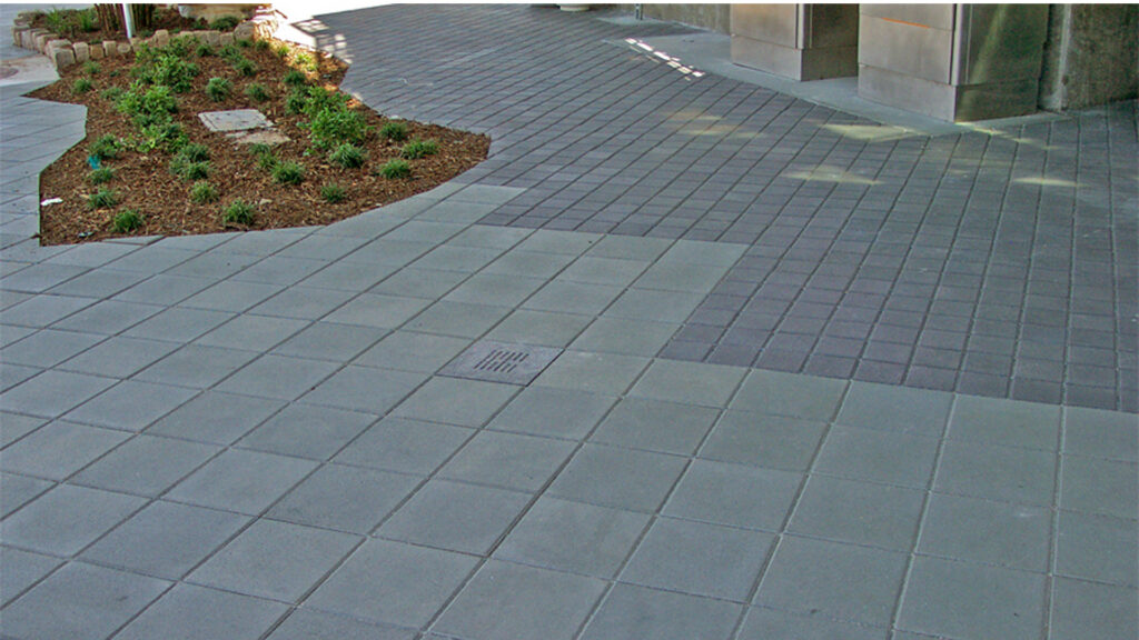 Paving installation featuring French Gray (special order) 12x12 square pavers in the foreground, transitioning into a contrasting section of Charcoal 6x6 pavers. The layout includes a centered drain and curves gently around a landscaped planting area.