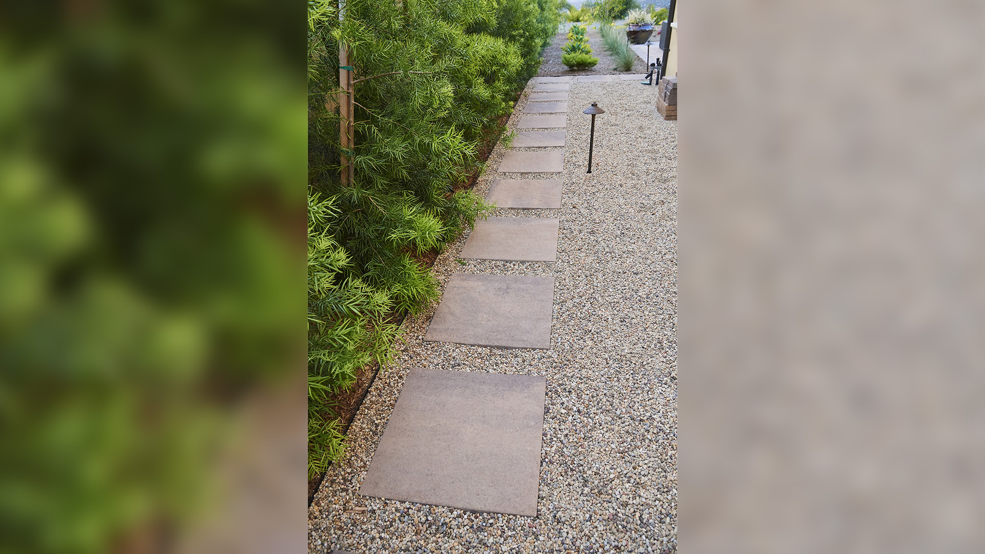 Narrow garden walkway featuring Adobe-Copper-Mocha Pavilion II stepstones set in a straight line over a bed of decorative gravel. The path is bordered by lush green foliage on one side and a modern home exterior on the other, with low landscape lighting providing subtle illumination.