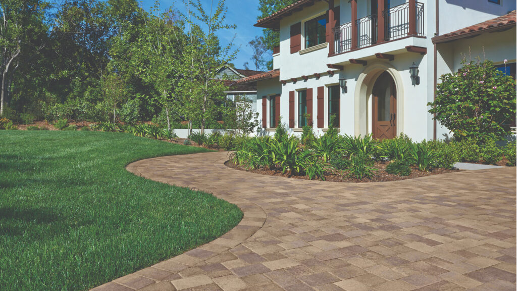 Curved driveway and walkway paved with Sand-Stone-Mocha Antique Cobble I & II arranged in an “I” pattern, leading up to a white Mediterranean-style home. The border features Sand-Stone-Mocha Antique Cobble I laid in a soldier course for definition. Lush green lawn and manicured landscaping frame the pathway.