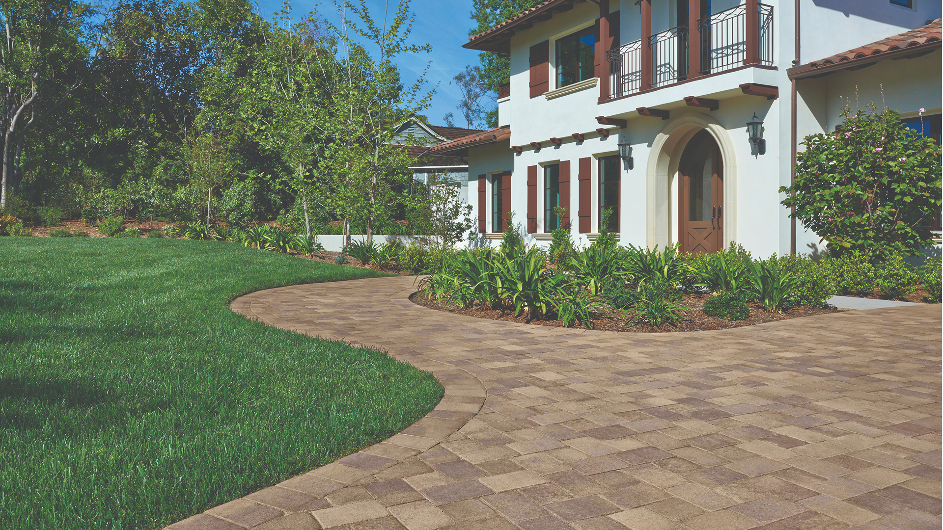 Curved driveway and walkway paved with Sand-Stone-Mocha Antique Cobble I & II arranged in an “I” pattern, leading up to a white Mediterranean-style home. The border features Sand-Stone-Mocha Antique Cobble I laid in a soldier course for definition. Lush green lawn and manicured landscaping frame the pathway.