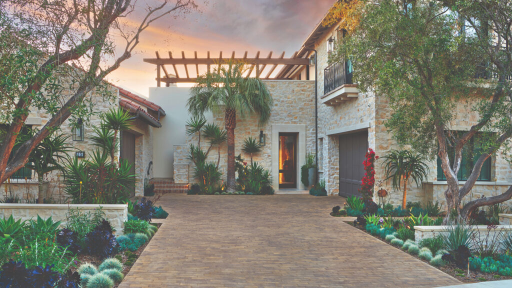 Residential driveway featuring Aqualina paving stones in Blended Sand Stone Mocha Blend, surrounded by Mediterranean-style landscaping with palm trees, succulents, and a stone-clad home at sunset.