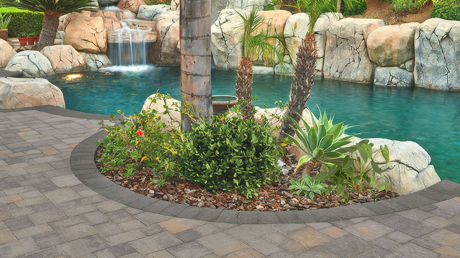 Tropical backyard poolscape featuring Slate Stone pavers in Dark Gray-Copper-Charcoal (special order) installed in a random ashlar pattern. The curved border is made of Slate 6x9 pavers in solid Charcoal. The design surrounds a vibrant planting bed with tropical greenery and palm trees, set beside a natural rock waterfall flowing into a turquoise pool.