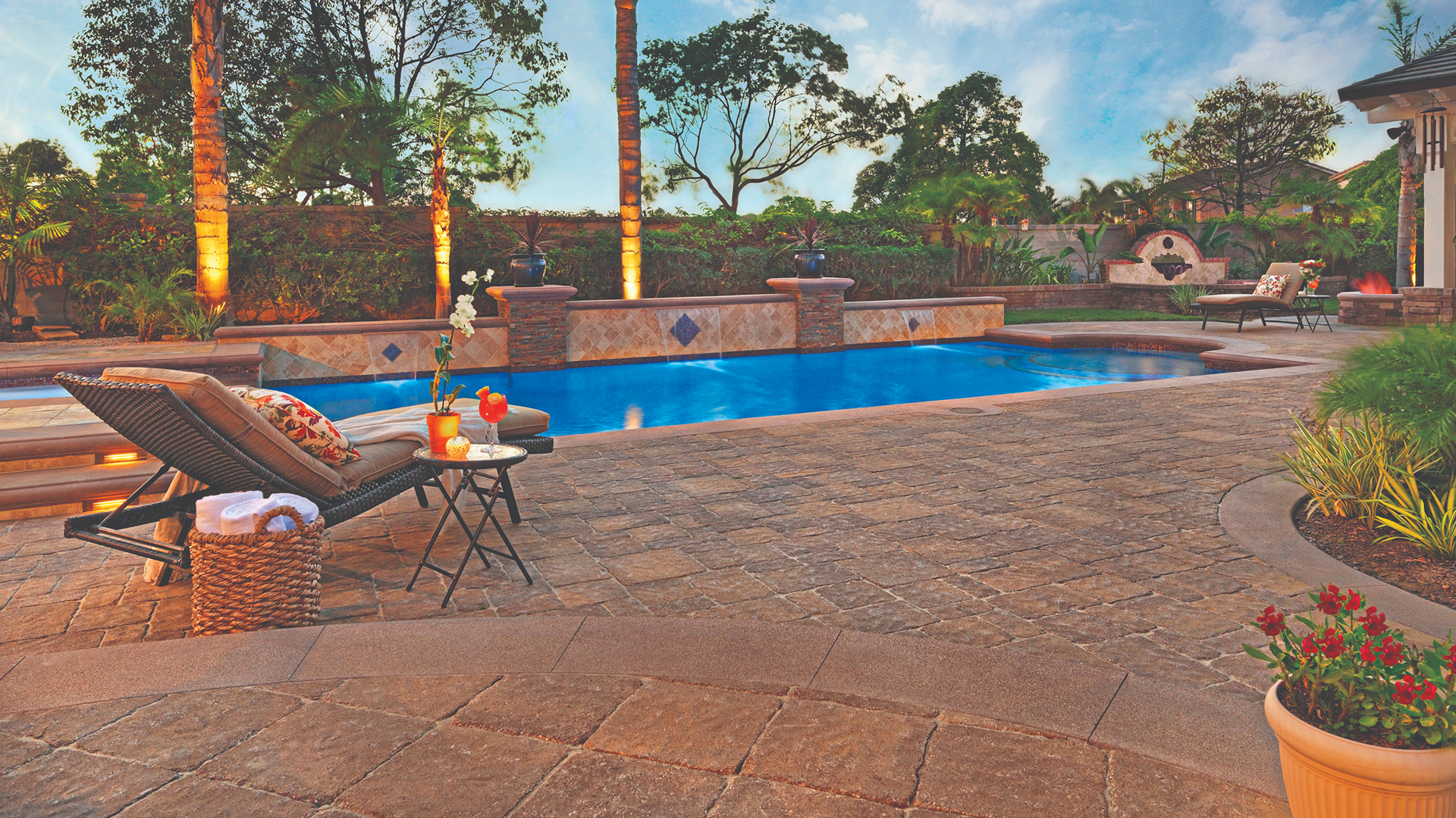 Backyard poolside patio featuring Sand Stone Mocha Estate Cobble I & II paving stones in a random pattern. A lounge chair and side tables are set near a spa-style pool with water features and lush landscaping in the background.