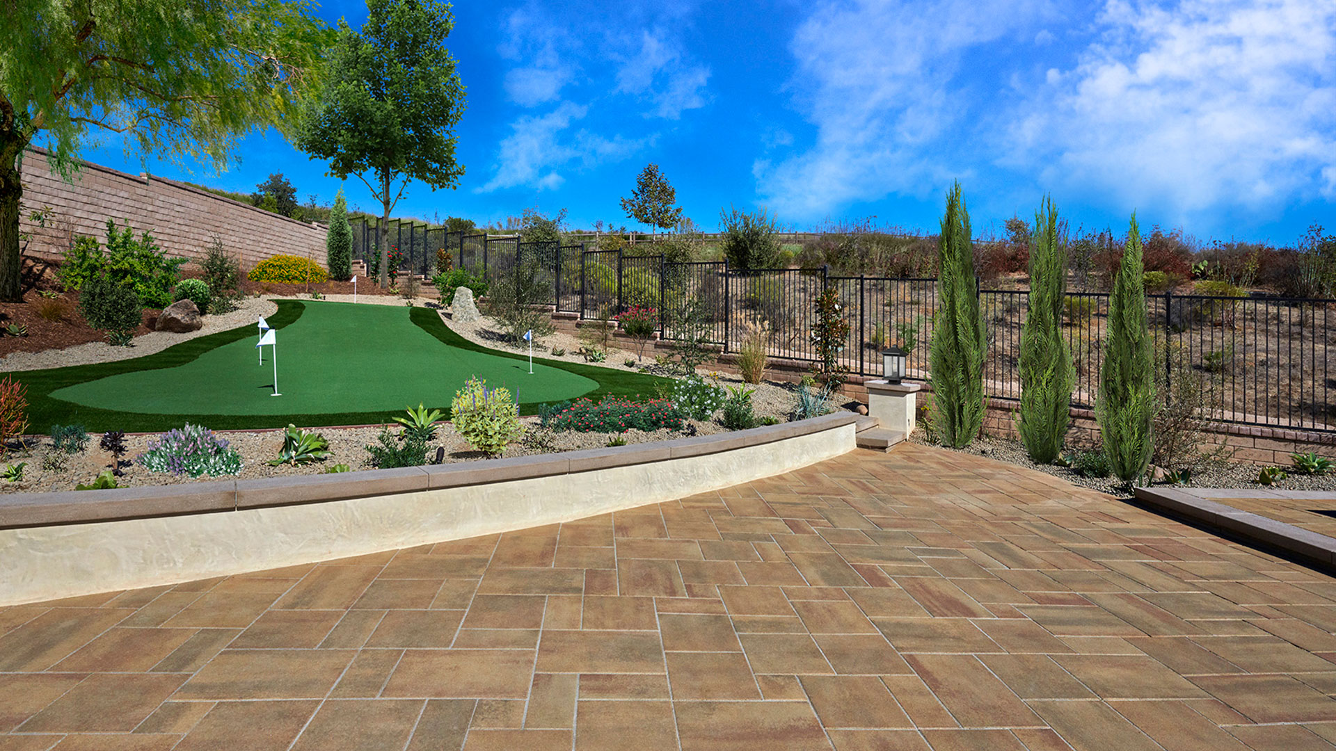 Blended Adobe-Copper-Mocha Paseo I & II paving stones installed in an ashlar pattern. The patio overlooks a putting green and desert-style landscaping, with cypress trees, flowering plants, and a view of a fenced hillside under a bright blue sky.
