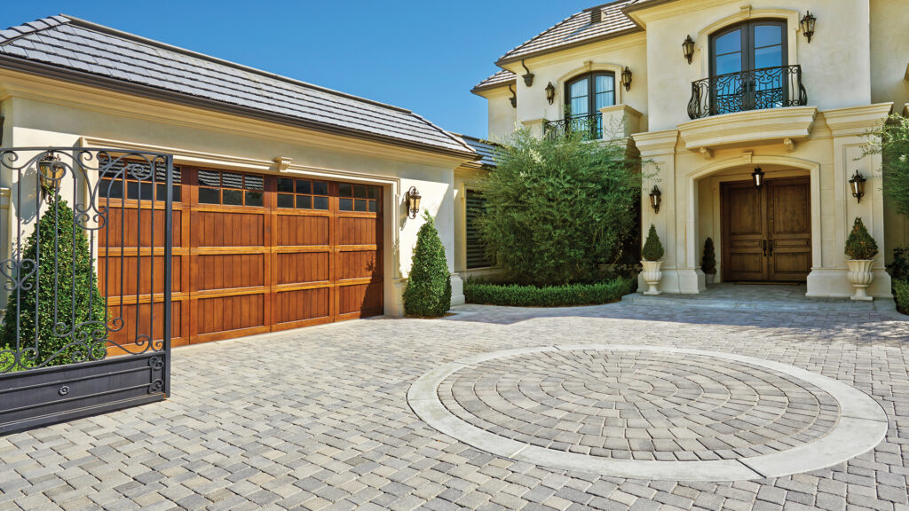 Elegant residential driveway featuring Gray-Charcoal Antique Cobble I and II (tumbled) paving stones installed in an “I” pattern. At the center is a decorative circle made of matching Gray-Charcoal Antique Cobble Circle (tumbled) stones. The home includes a wooden garage door, grand double-door entry, wrought iron accents, and neatly manicured landscaping, creating a sophisticated and timeless curb appeal.