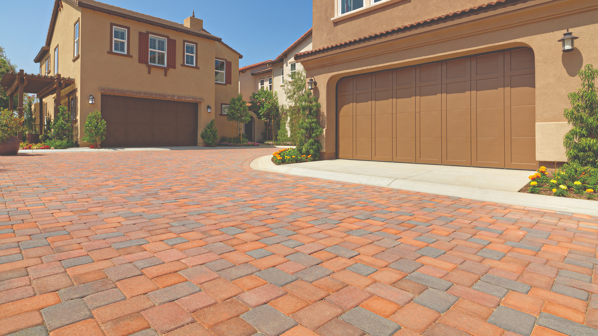 Residential community driveway featuring Antique Cobble I & II in a 50/50 blend of Terracotta-Brown and Red-Brown-Charcoal, installed in an 'I' pattern, complementing the warm tones of the surrounding stucco homes and brown garage doors.