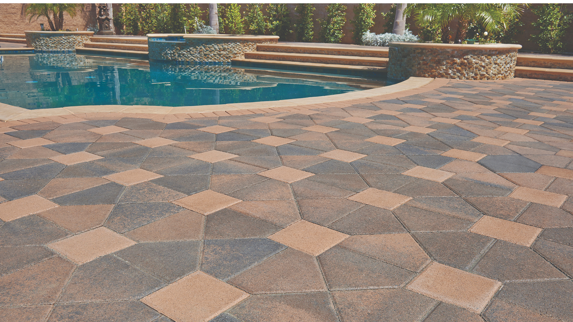 Poolside patio featuring Cream-Brown-Charcoal Bishops Hat pavers paired with Cream 8x8 pavers, arranged in a Venetian parquet pattern. The design is framed with a clean Cream Holland paver border. The scene includes curved pool coping and a raised spa with stacked stone facing, set against a backdrop of natural landscaping and steps leading to a higher terrace.