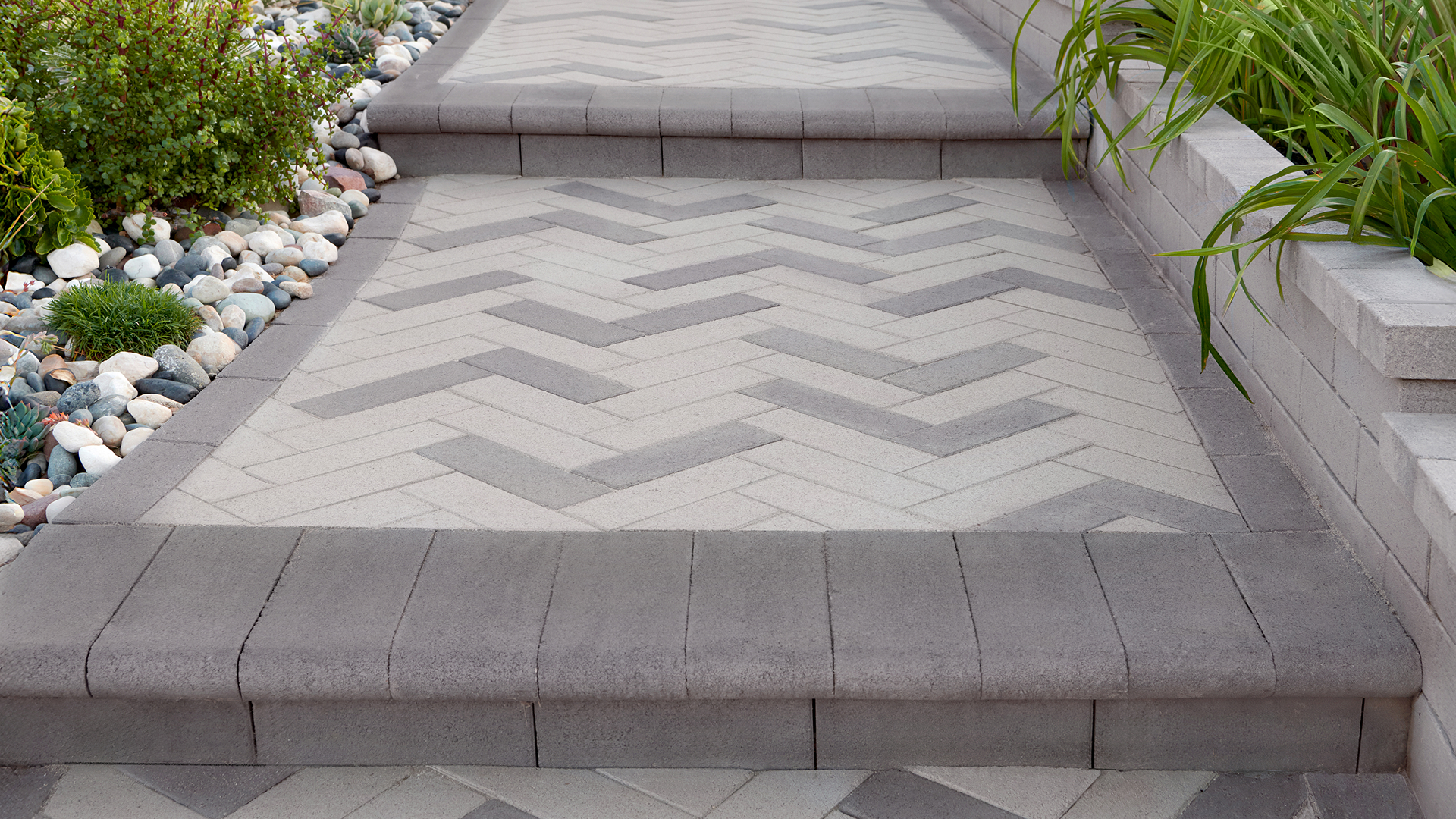 Modern garden walkway and steps featuring a herringbone pattern made from 80mm 4x12 pavers in a field blend of solid Gray and solid Pewter. The border and stair treads are finished with Charcoal 6x14 Bullnose pavers. The path is flanked by decorative river rocks, low green shrubs, and a raised planter wall, creating a clean and contemporary landscape design.