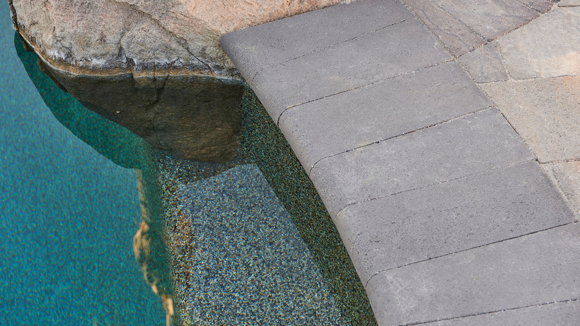 Close-up view of a pool edge finished with 6x14 Bullnose coping in solid Charcoal. The surrounding patio surface features Slate Stone pavers in Dark Gray-Copper-Charcoal arranged in a random ashlar pattern, with a Slate 6x9 solid Charcoal border. Natural rock elements blend into the pool’s curved edge, creating a seamless transition between hardscape and water.
