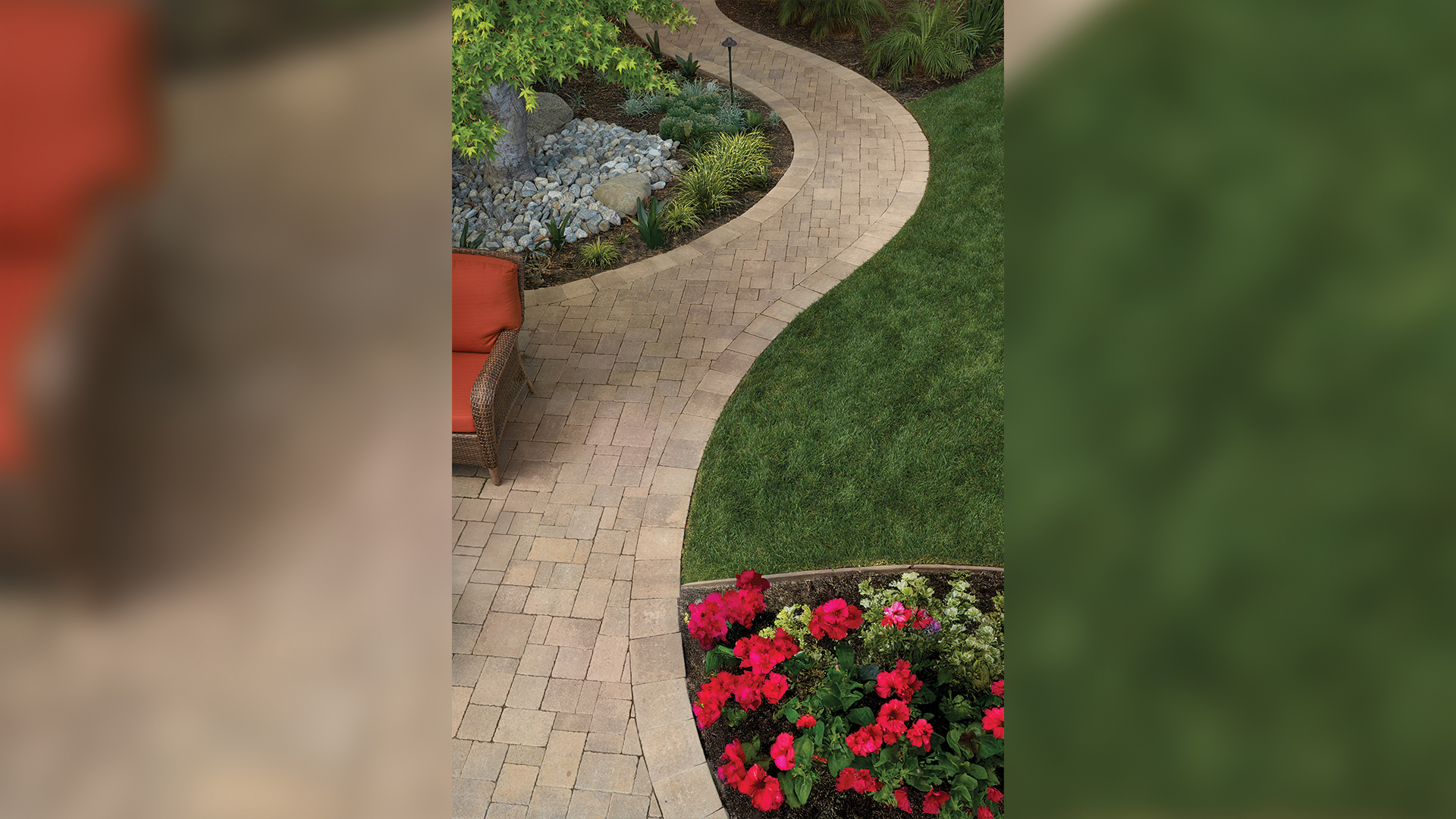 Curving garden pathway and patio area paved with Sand-Stone-Mocha Castle Cobble I & II (distressed) stones, arranged in a random ashlar pattern. The border features Sand-Stone-Mocha Castle Cobble II in a running bond layout. The scene includes a wicker chair with orange cushions, a vibrant flower bed with red and pink blooms, lush green grass, and surrounding landscape with decorative rocks and plants beneath a tree.