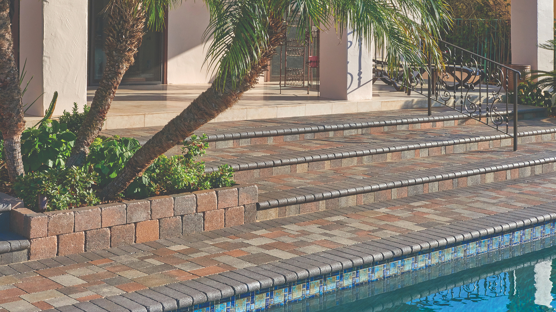 Outdoor poolside area featuring Castle Cobble I and II pavers with a distressed texture, installed in a 50/50 field mix of Cream-Brown-Charcoal and Gray-Moss-Charcoal in an ashlar pattern. Steps and pool coping are finished with solid Charcoal Bullnose. A raised planter near the palm tree is built with Rustic Wall blocks in Cream-Brown-Charcoal.