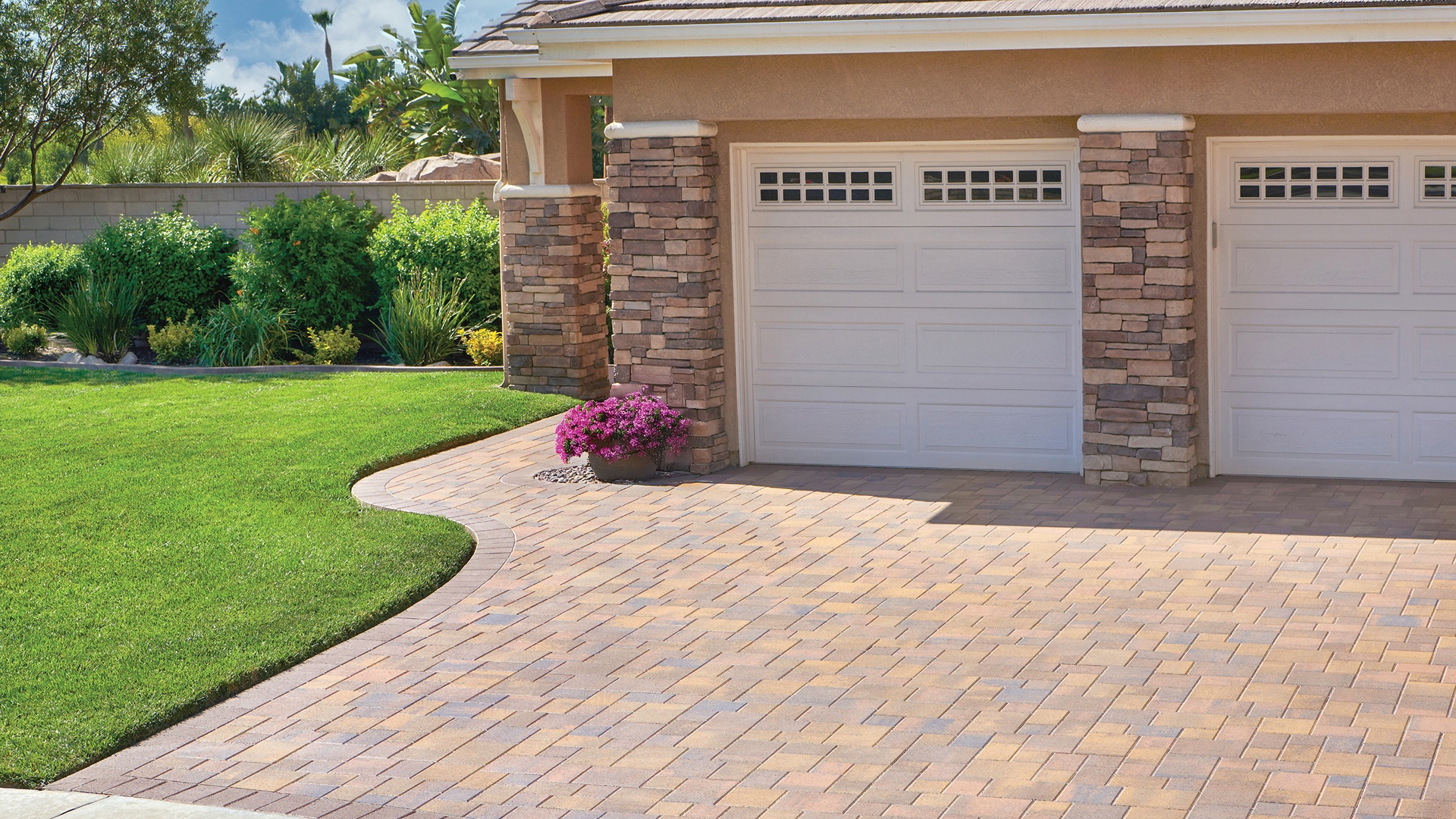 Residential driveway paved with Courtyard Stone in a 50/50 field blend of Cream-Brown-Charcoal and Sand-Stone-Mocha colors, arranged in a random pattern. The border features solid Mocha Courtyard 6x9 pavers installed in a soldier course. The driveway leads to a double garage with white paneled doors and stacked stone columns, bordered by lush green grass, colorful landscaping, and a potted plant with vibrant pink flowers.