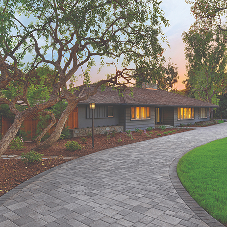 Curved driveway paved with Courtyard Combo in Sand-Stone-Mocha blend, installed in a random ashlar pattern. The setting features a single-story home with dark siding, mature trees, and a manicured lawn under a glowing sunset sky.