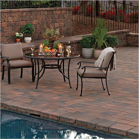 looking over an outdoor pool you see a couple of chairs and table on the pool deck. a tiered wall is behind the table and chairs. the tiered wall has some throw pillows and blanket for seating. wall is built with blended cream brown charcoal stonewall two. the pool deck in covered in blended cream brown charcoal courtyard stone paving stones installed in an ashlar pattern.