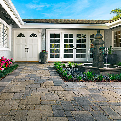 Heartland paving stones in a Gray-Moss-Charcoal blend installed in a random ashlar pattern, with a 6x12 Gray-Charcoal border laid in a soldier course (special order). The elegant entryway features a tiered fountain, manicured plantings, and a white double-door entrance with surrounding French doors.
