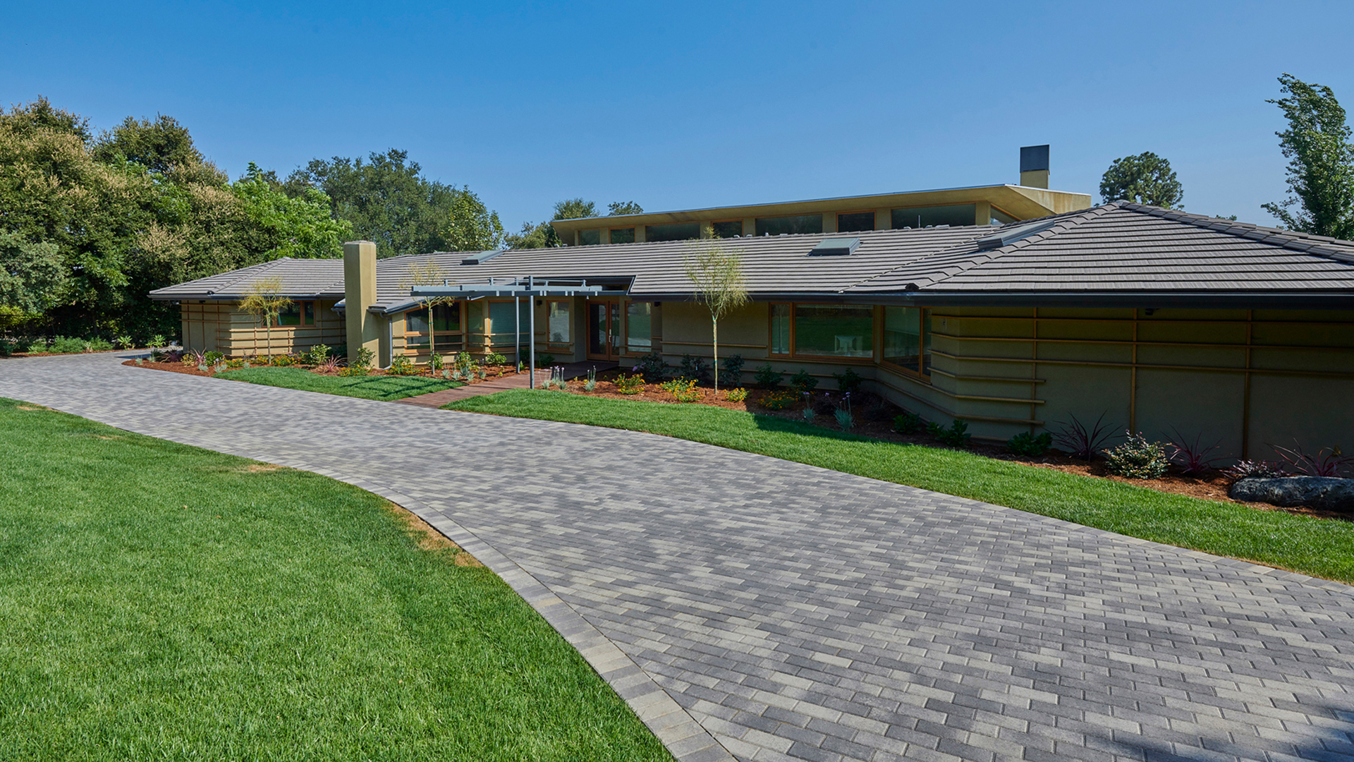 Modern residential driveway paved with Holland pavers in a Gray-Charcoal color blend, installed in a running bond pattern. The border is made of matching Holland Gray-Charcoal pavers set in a soldier course. The driveway curves past a contemporary single-story home with a landscaped front yard, manicured lawn, and clean architectural lines.