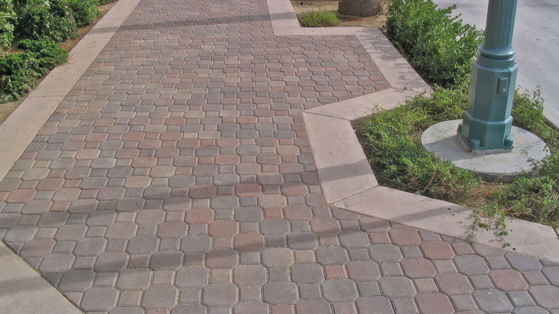 sidewalk with posts, tree, and street on the right. Bushes on the left. sidewalk covered in blended terracotta brown keylock paving stones. sidewalk border is cream brown keylock paving stones.