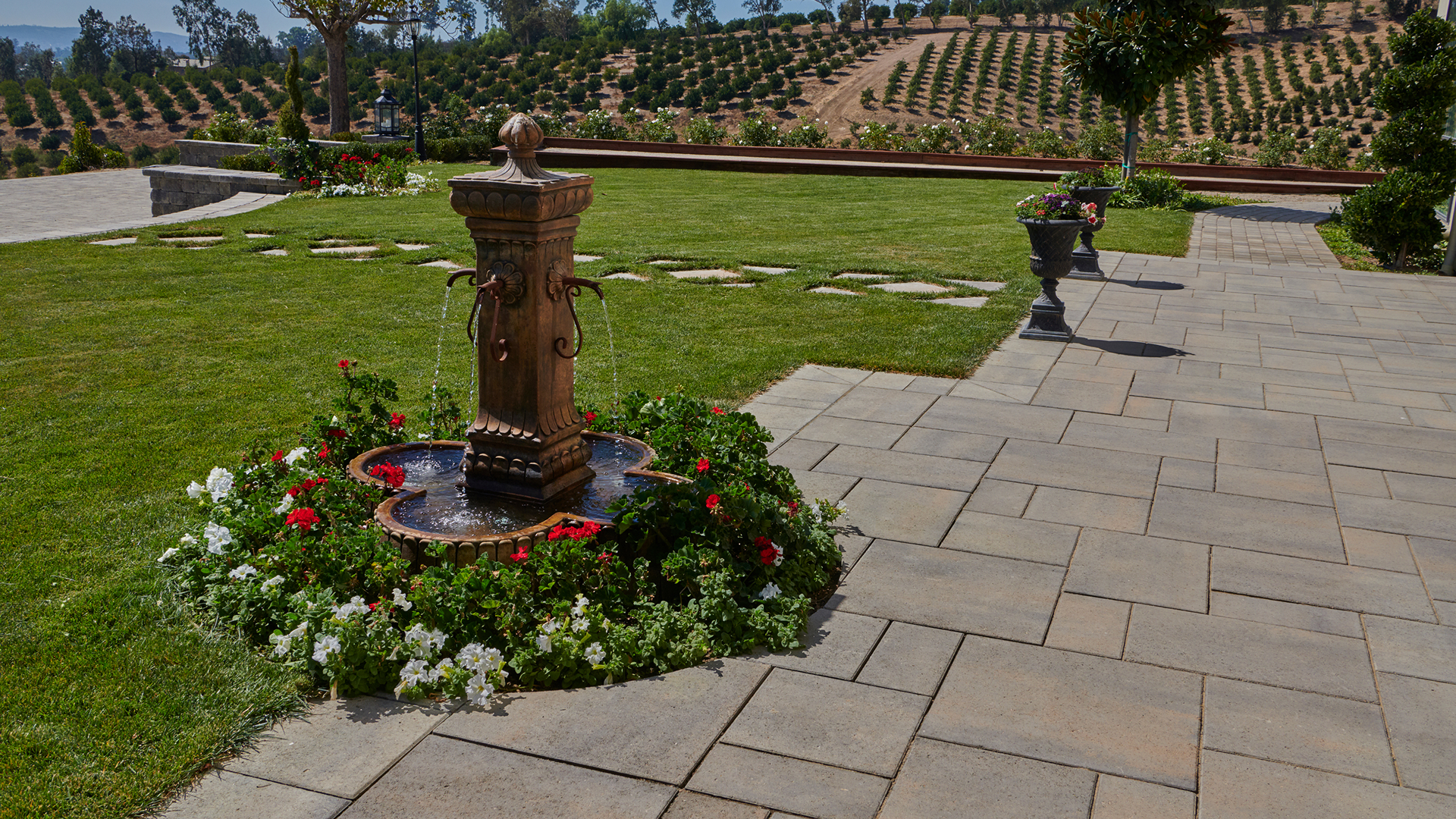 Elegant outdoor space featuring Paseo I & II pavers in Dark Gray-Copper-Charcoal, installed in a random ashlar pattern. The scene includes a bronze decorative fountain surrounded by vibrant red and white flowers, a manicured lawn, ornamental planters, and scenic vineyard-style hills in the background. The classic paver layout complements the formal garden and expansive landscape design.
