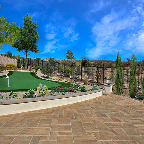Blended Adobe-Copper-Mocha Paseo I & II paving stones installed in an ashlar pattern. The patio overlooks a putting green and desert-style landscaping, with cypress trees, flowering plants, and a view of a fenced hillside under a bright blue sky.