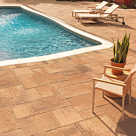 Poolside patio featuring Pavilion I and II pavers in Adobe-Copper-Mocha, installed in a random ashlar pattern. The curved pool deck is bordered with Slate Stone 6x9 pavers in Sand-Stone-Mocha. The scene includes modern lounge chairs and a potted plant under bright sunlight, with water gently cascading into the pool.