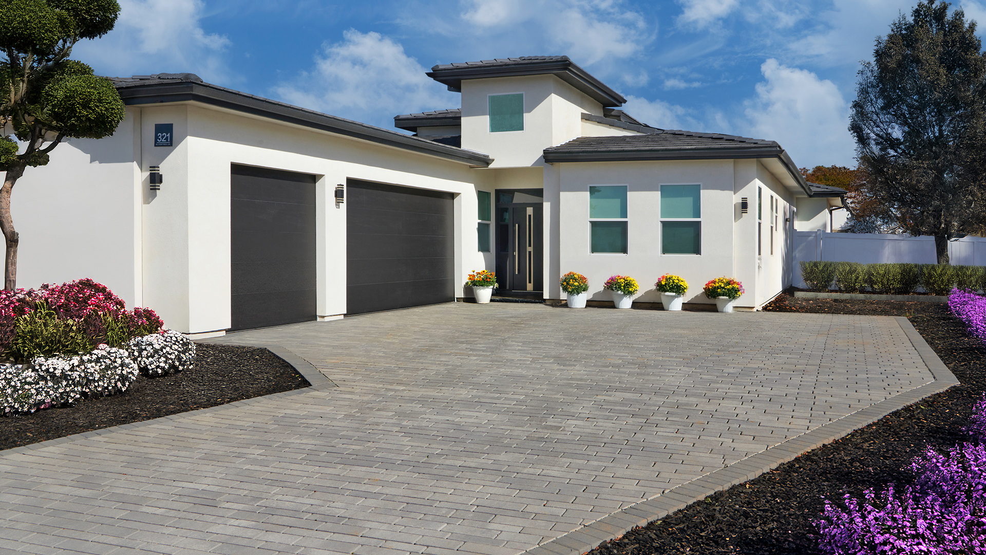 Modern front driveway featuring 100mm Permeable Aqualina pavers in Dark Gray-Pewter-Charcoal installed in a random runner pattern. The border is composed of 60mm Holland pavers in Solid Charcoal laid in a soldier course. The scene includes a white contemporary house with black garage doors, flower beds with colorful blooms, manicured shrubs, and a bright blue sky with scattered clouds.