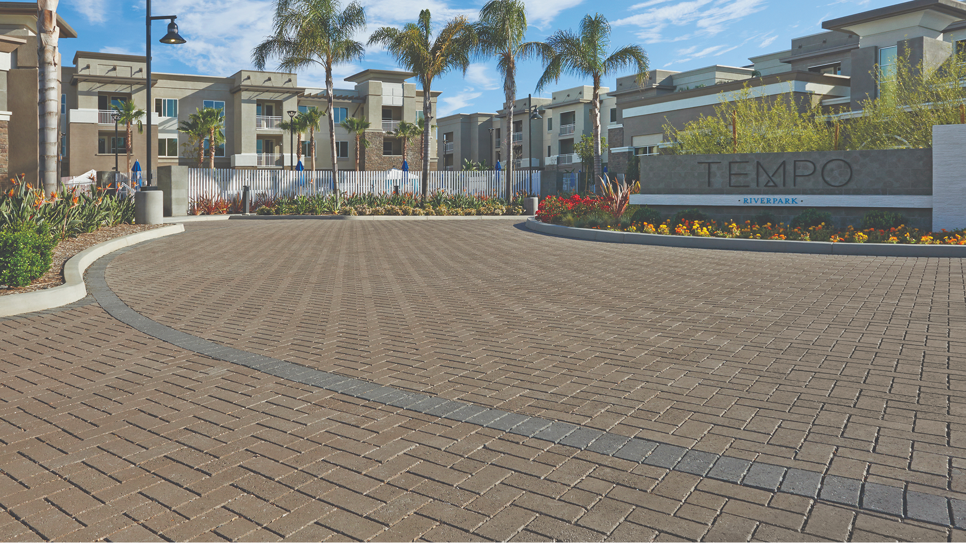Entryway to a commercial housing development paved in Solid Brown permeable Holland paving stones arranged in a herringbone pattern, with a curved band of Solid Charcoal permeable Holland pavers for contrast. Modern apartment buildings and landscaped gardens surround the entrance.