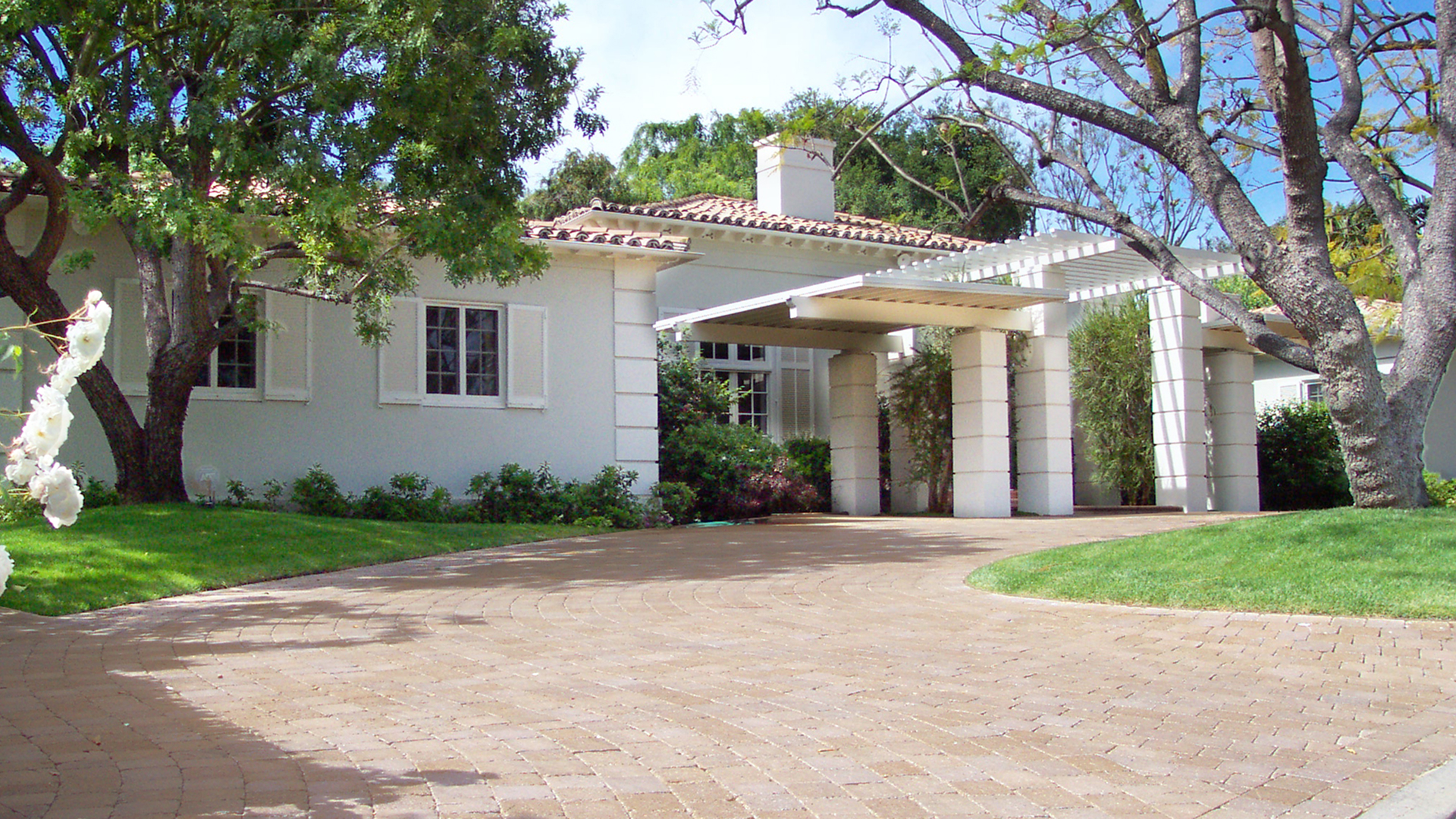Curved driveway paved with Sand-Stone Tumbled Quarry Cobble in a running bond pattern, surrounded by manicured landscaping, flowering shrubs, and a green lawn, leading to a home with a white columned portico entrance.