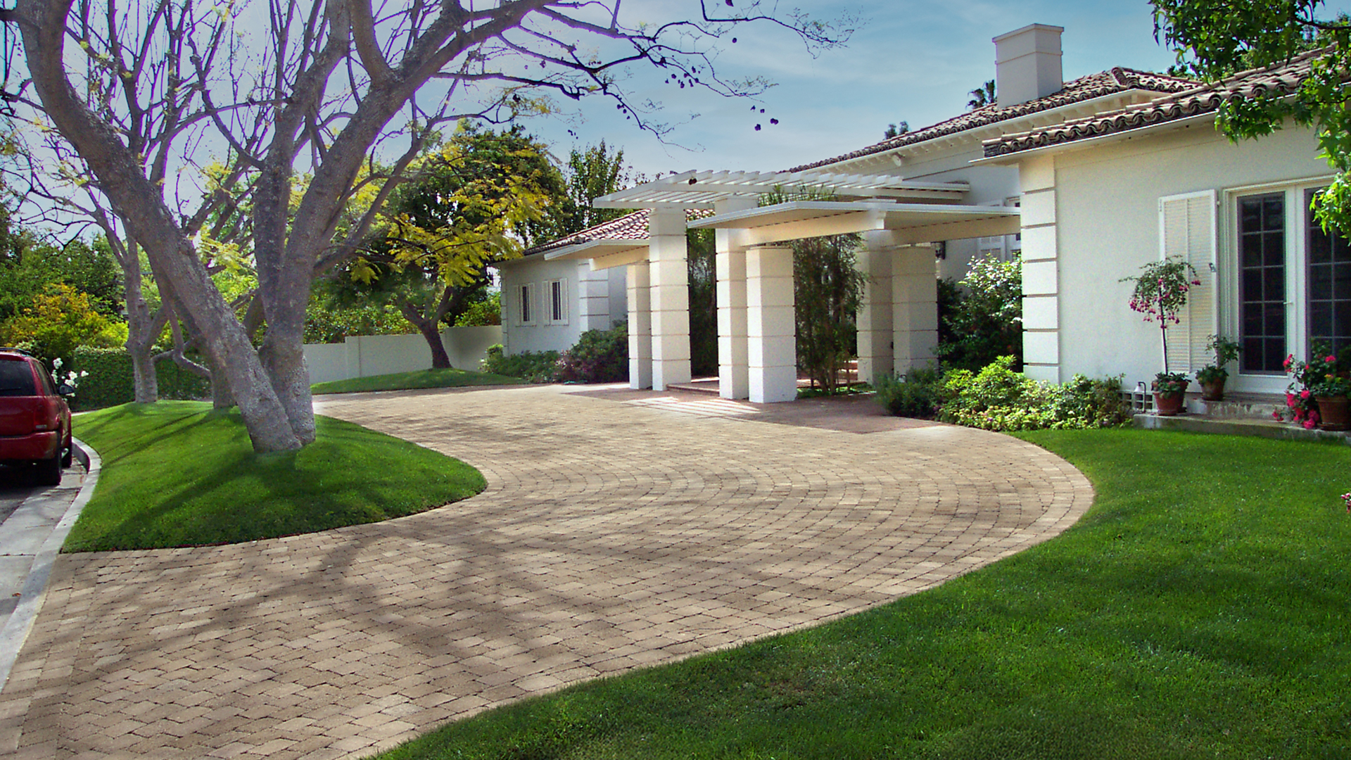 Driveway paved with Sand-Stone Tumbled Quarry Cobble pavers in a running bond pattern, curving around a green lawn with mature trees and leading to a white-columned portico entrance of a residential home.