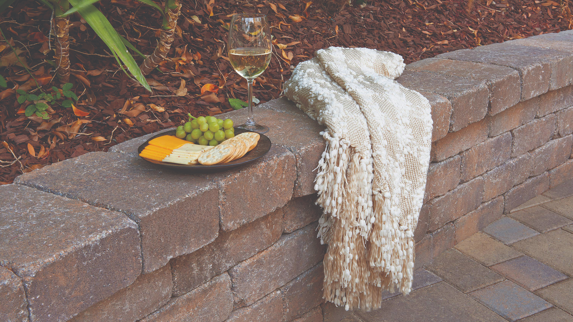 Tuscan Rustic Wall used as a seating wall with a cozy cream-colored throw draped over the edge, beside a plate of cheese, crackers, and grapes with a glass of white wine, set against a mulched garden backdrop.