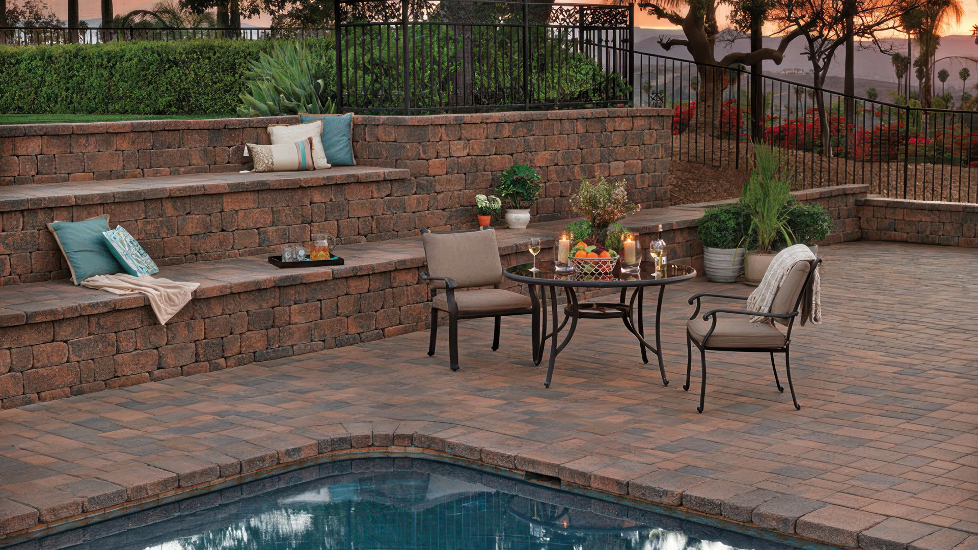 looking over an outdoor pool you see a couple of chairs and table on the pool deck. a tiered wall is behind the table and chairs. the tiered wall has some throw pillows and blanket for seating. wall is built with blended cream brown charcoal stonewall two. the pool deck in covered in blended cream brown charcoal courtyard stone paving stones installed in an ashlar pattern.