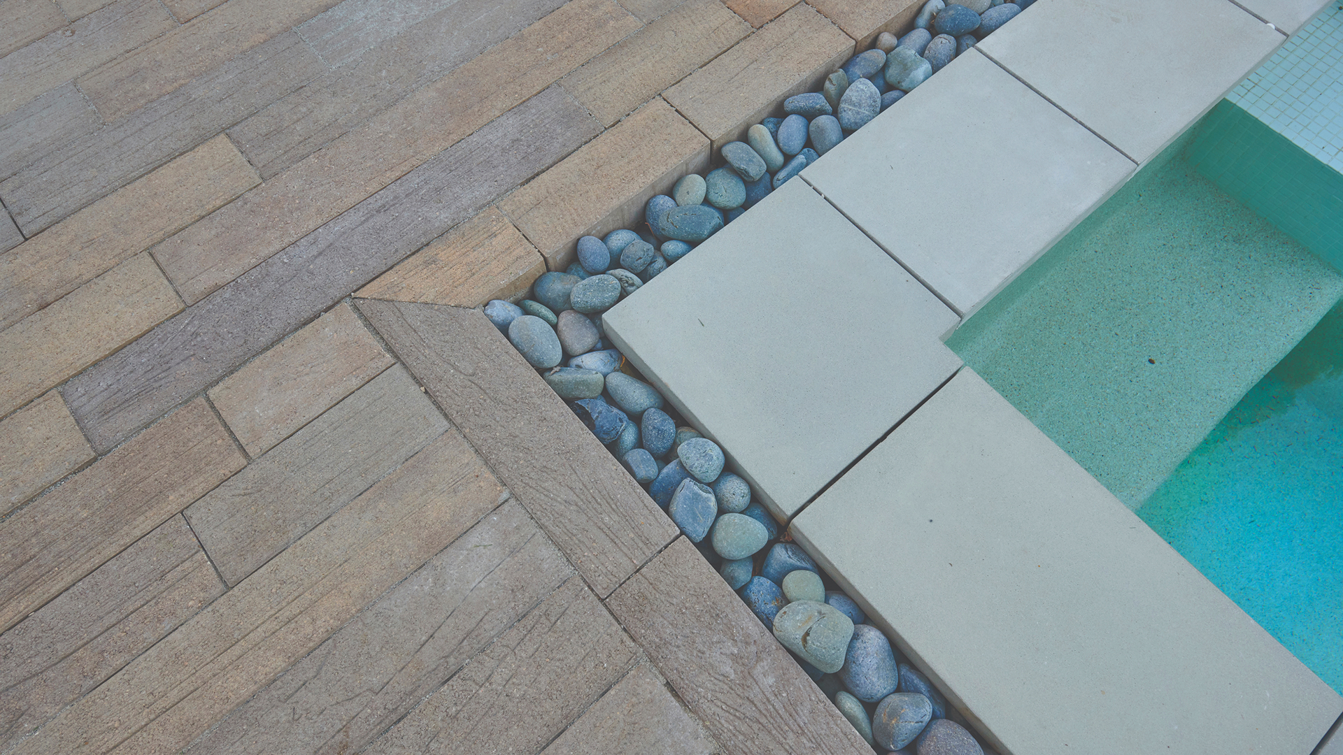 Close-up of a poolside deck featuring 70mm Timberline pavers in a Stone-Adobe-Mocha color blend, installed in a random running bond pattern. The paving transitions to smooth concrete pool coping with a decorative strip of blue river rocks in between, creating a clean and natural contrast alongside the aqua-toned water of the pool.