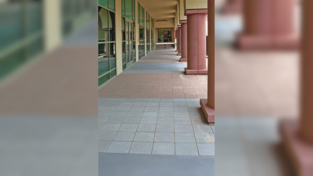 Covered walkway along a commercial or institutional building with large glass windows and red columns. The ground is paved with alternating sections of Gray and Cream 12x12 square pavers, installed in a stack bond pattern. The alternating colors create a subtle visual rhythm along the path.
