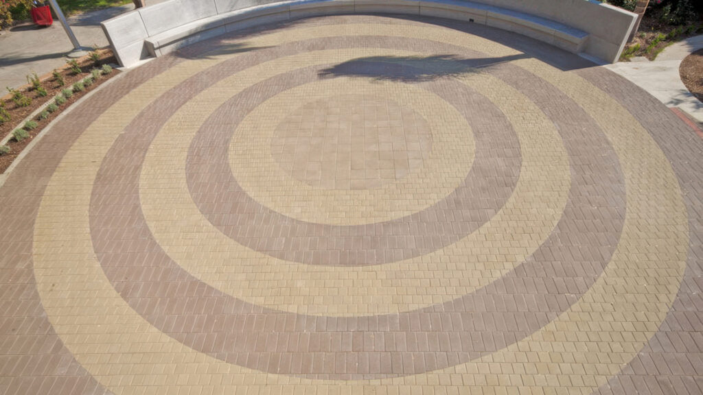 Circular plaza design with alternating ring patterns made from Red 6x6 square pavers in a running bond layout, followed by Mocha 6x12 and Moss 6x6 square pavers forming concentric bands. At the center is a large 12x12 square area arranged in a stack bond pattern. The space is framed by curved concrete benches and landscaped planting beds, creating a modern and inviting gathering area.