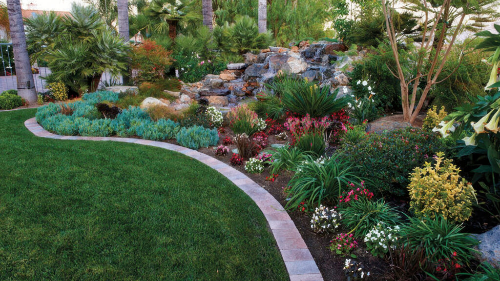 Edging features a single curved band of Cream-Brown-Charcoal Slate Stone 6x9 (distressed), separating a manicured lawn from a vibrant garden bed filled with colorful plants and a rock waterfall feature.