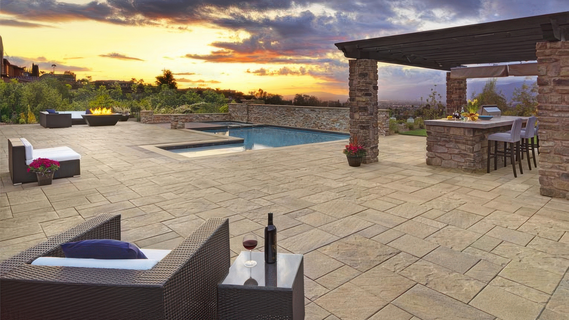 Pavilion I and II pavers in Adobe-Copper-Mocha installed in a random ashlar pattern surround a modern outdoor living space featuring a pool, fire pit, and bar area. The paver border is Slate 6x9 in Cream-Brown-Charcoal, laid in a soldier course. Stylish wicker seating and a panoramic sunset complete the elegant backyard setting.