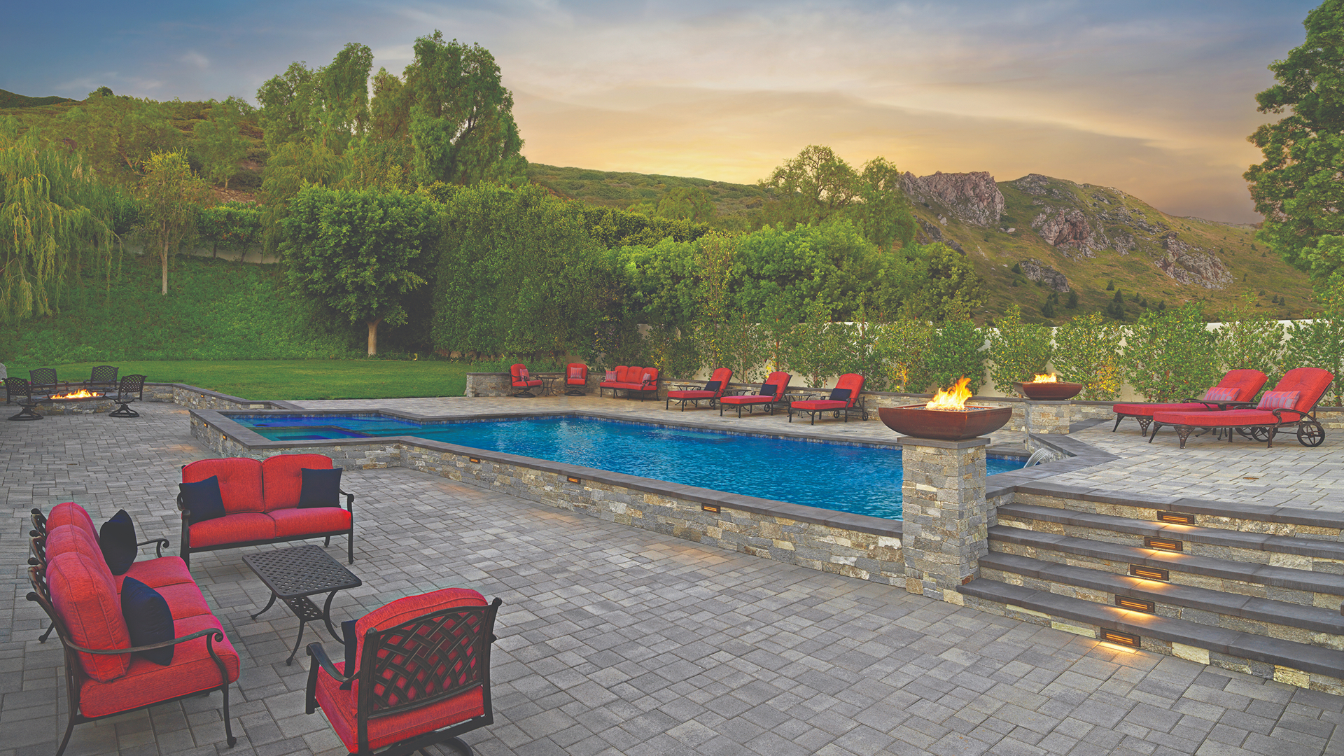 Spacious backyard featuring Courtyard Stone pavers in Dark Gray-Pewter-Charcoal, arranged in a random ashlar pattern around a modern rectangular pool. The scene includes red cushioned lounge chairs, a fire feature at the poolside, and built-in stair lighting, all set against a lush hillside backdrop at sunset.