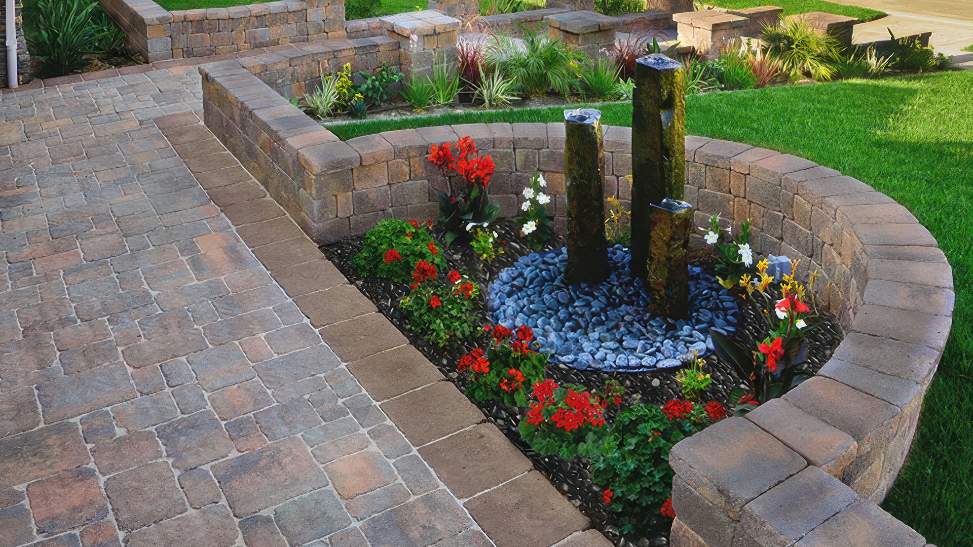Outdoor landscape featuring Tuscan Stonewall® II blocks and caps installed in a random ashlar pattern forming raised planter beds and seating walls. Paving area showcases Estate Cobble I & II (tumbled) pavers in Tuscan with a random ashlar layout. A solid Mocha (special order) Estate Cobble II tumbled border defines the edge. A circular fountain with three stone pillars is surrounded by vibrant red, white, and yellow flowers.