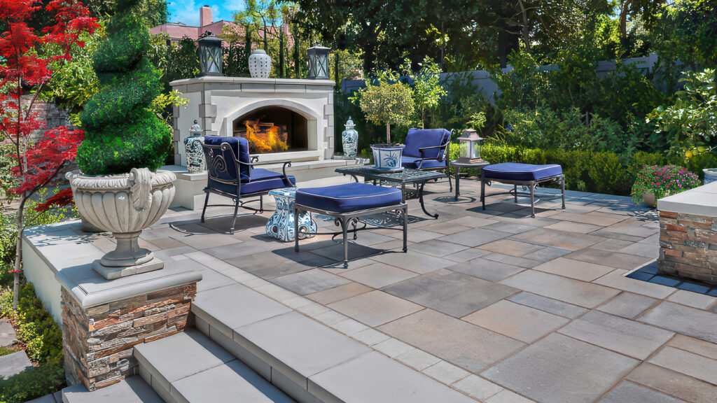 Outdoor patio featuring Paseo I & II pavers in Dark Gray-Copper-Charcoal, arranged in a random ashlar pattern. Elegant seating surrounds a built-in fireplace, all set within a lush garden landscape.