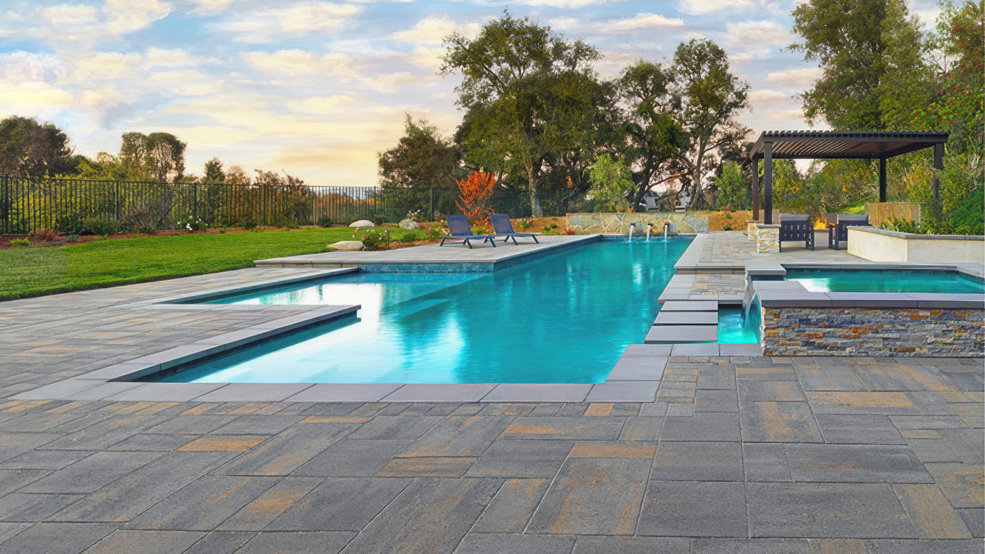 Modern backyard pool and spa area featuring Paseo I & II pavers in Dark Gray-Copper-Charcoal, installed in a random ashlar pattern. A narrow border of Courtyard 6x9 pavers in the same color runs along the pool coping. The space includes lounge chairs, a pergola-covered seating area, and landscaped greenery under a soft sunset sky.