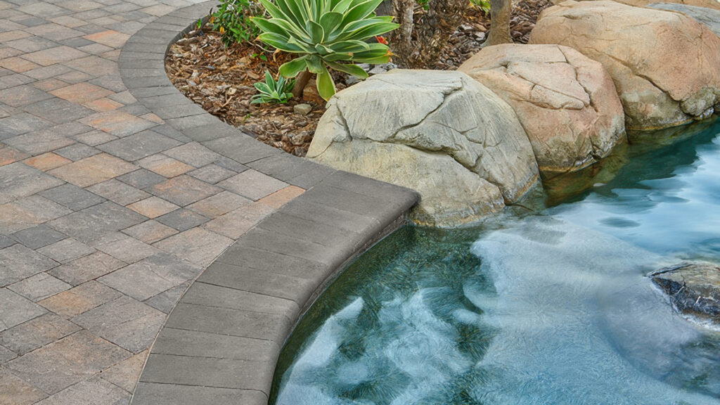 Close-up view of a poolside with Slate Stone pavers in Dark Gray-Copper-Charcoal installed in a random ashlar pattern. The layout includes a border of solid Charcoal 6x9 Slate Stone and a coping edge of solid Charcoal 6x14 bullnose. Natural boulders and lush plants accent the water's edge.