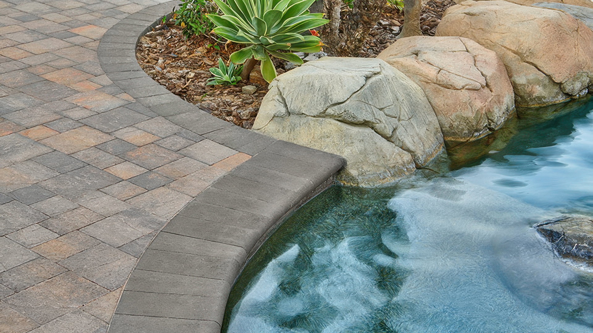Close-up view of a poolside with Slate Stone pavers in Dark Gray-Copper-Charcoal installed in a random ashlar pattern. The layout includes a border of solid Charcoal 6x9 Slate Stone and a coping edge of solid Charcoal 6x14 bullnose. Natural boulders and lush plants accent the water's edge.