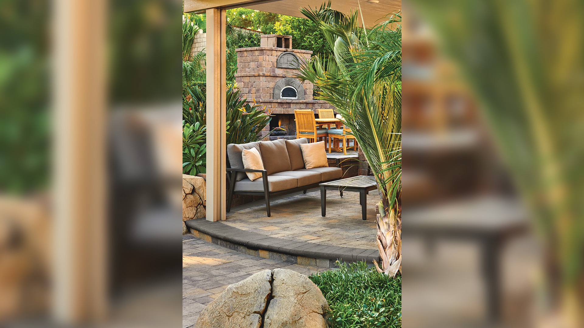 Covered patio area featuring Slate Stone pavers in a Dark Gray-Copper-Charcoal blend, installed in a random ashlar pattern. A raised seating area is bordered by a step finished with 6x14 Bullnose in solid Charcoal. A stone pizza oven and wooden dining set are visible in the lush, landscaped background.