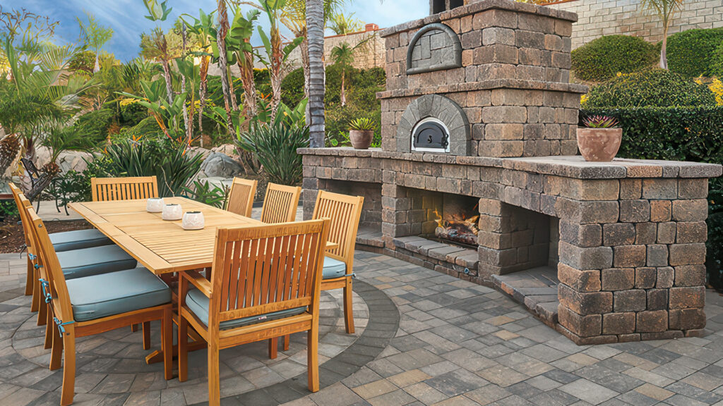 Elegant outdoor dining area featuring a custom-built Tuscan Stonewall® II fireplace constructed from block and cap. The patio is paved with Slate Stone pavers in a Dark Gray-Copper-Charcoal color, laid in a random ashlar pattern. Banding around the area is done with Slate 6x9 pavers in solid Charcoal, and a circular inlay uses Slate Circle pavers in Gray-Charcoal. Surrounded by lush landscaping and a wooden dining set.
