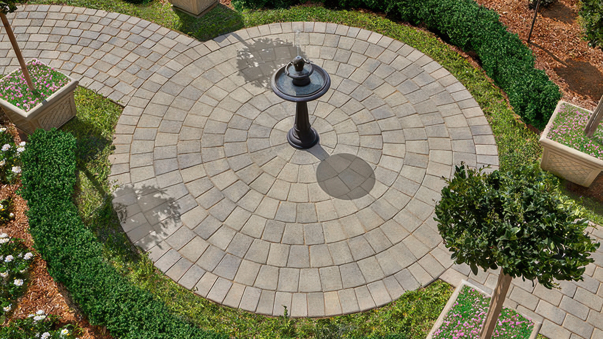 Circular patio features the Antique Cobble Circle package in Gray-Moss-Charcoal, centered around a black pedestal birdbath. Surrounding pathways use Antique Cobble I and II pavers laid in a random runner pattern, framed by manicured hedges, flower planters, and blooming garden beds.