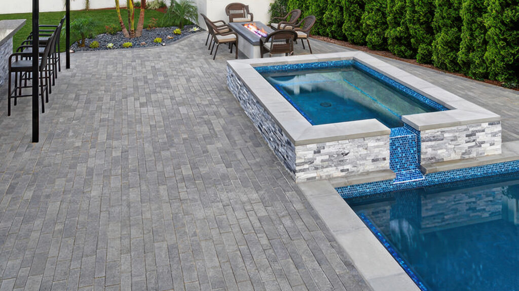 Modern backyard pool and spa area featuring Aqualina pavers in Dark Gray-Pewter-Charcoal, installed in a random runner pattern. The sleek design includes a raised spa with a cascading water feature into the pool, surrounded by a built-in bar area with seating and a fire table with additional chairs, all framed by manicured landscaping and evergreen privacy hedges.