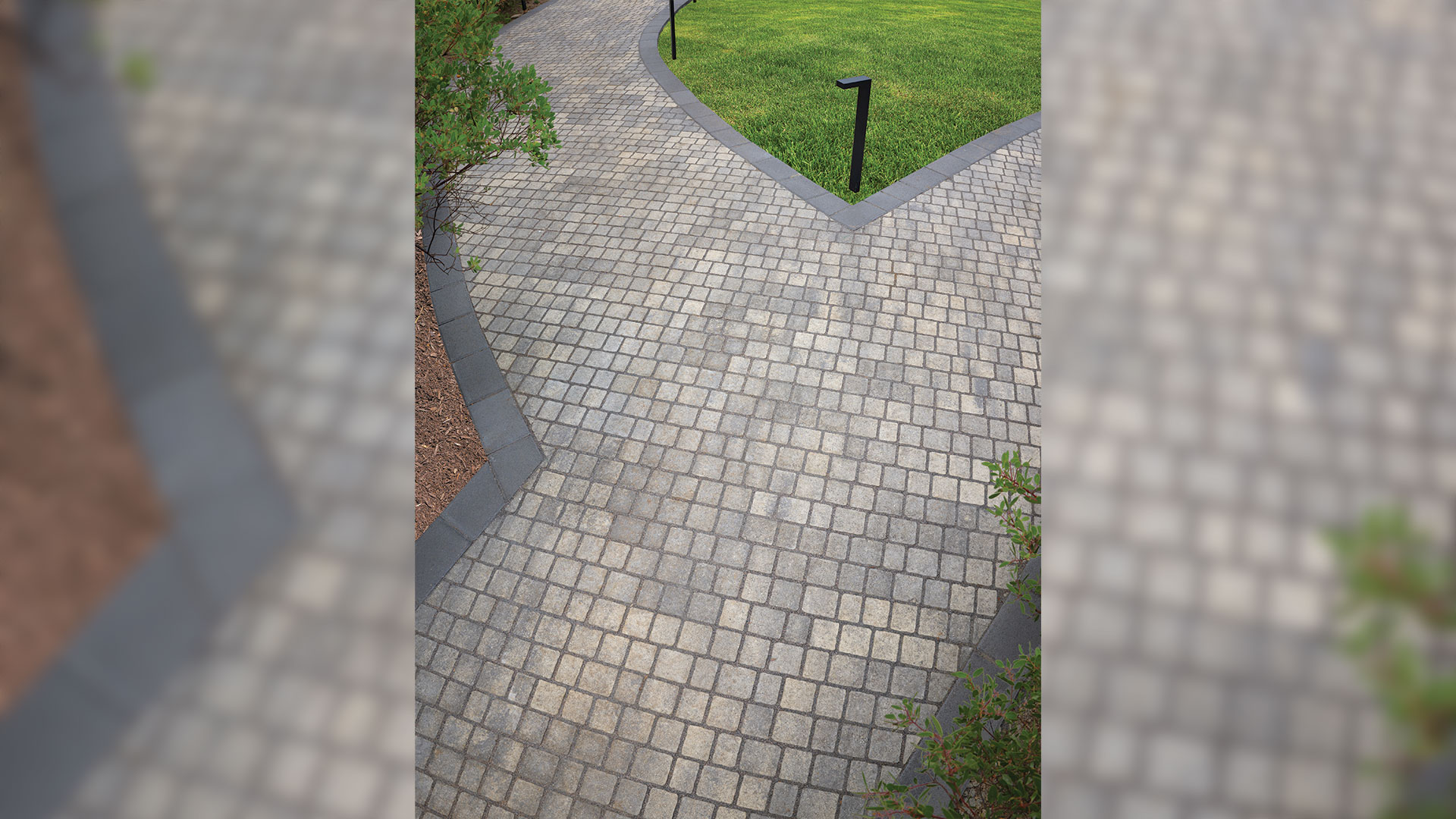 Curved garden walkway featuring 70mm Torino pavers in Gray-Charcoal installed in a random running bond pattern, bordered with Courtyard 6x9 pavers in Charcoal installed in a running bond pattern. The pathway winds through landscaped lawn and planting beds, with dark border accents defining the edges and highlighting the contrasting gray paver surface.