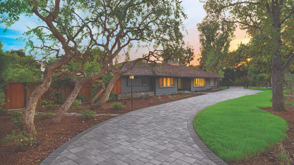 Curved driveway paved with Courtyard Combo in Sand-Stone-Mocha blend, installed in a random ashlar pattern. The setting features a single-story home with dark siding, mature trees, and a manicured lawn under a glowing sunset sky.
